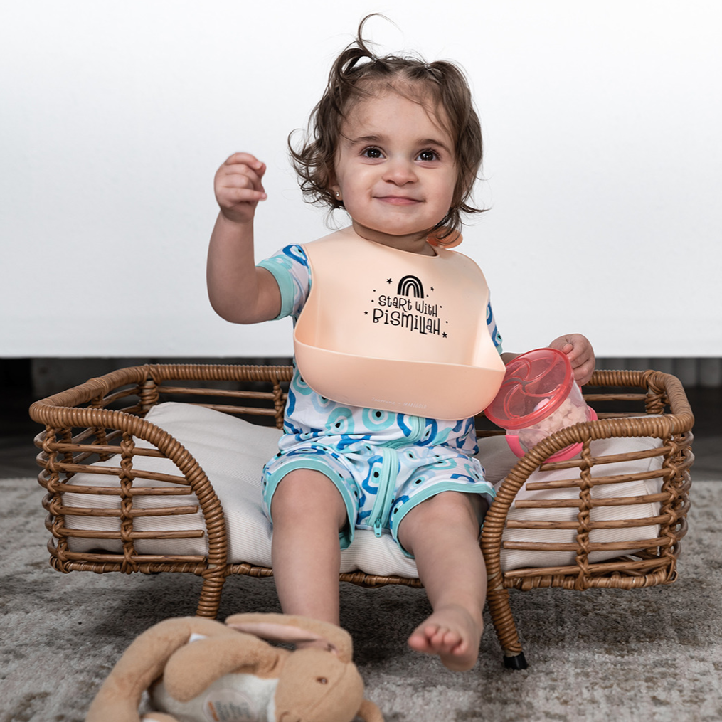 Baby sitting in small wicker bed wearing our Start With Bismillah silicone bib with flexible pouch. Wearing our evil eye shorty zippy