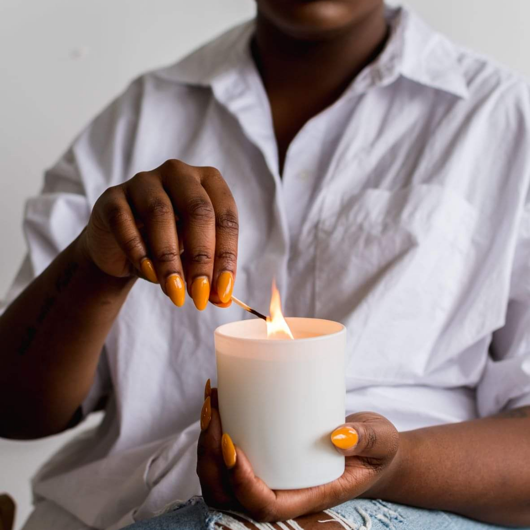 A woman in a white cotton shirt is lighting a luxury candle with a match. 