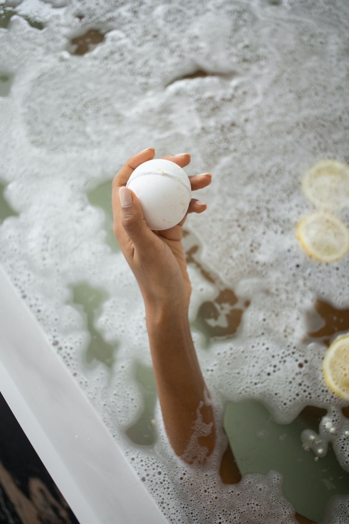 Woman holds a white bath bomb on top of the bath water. 