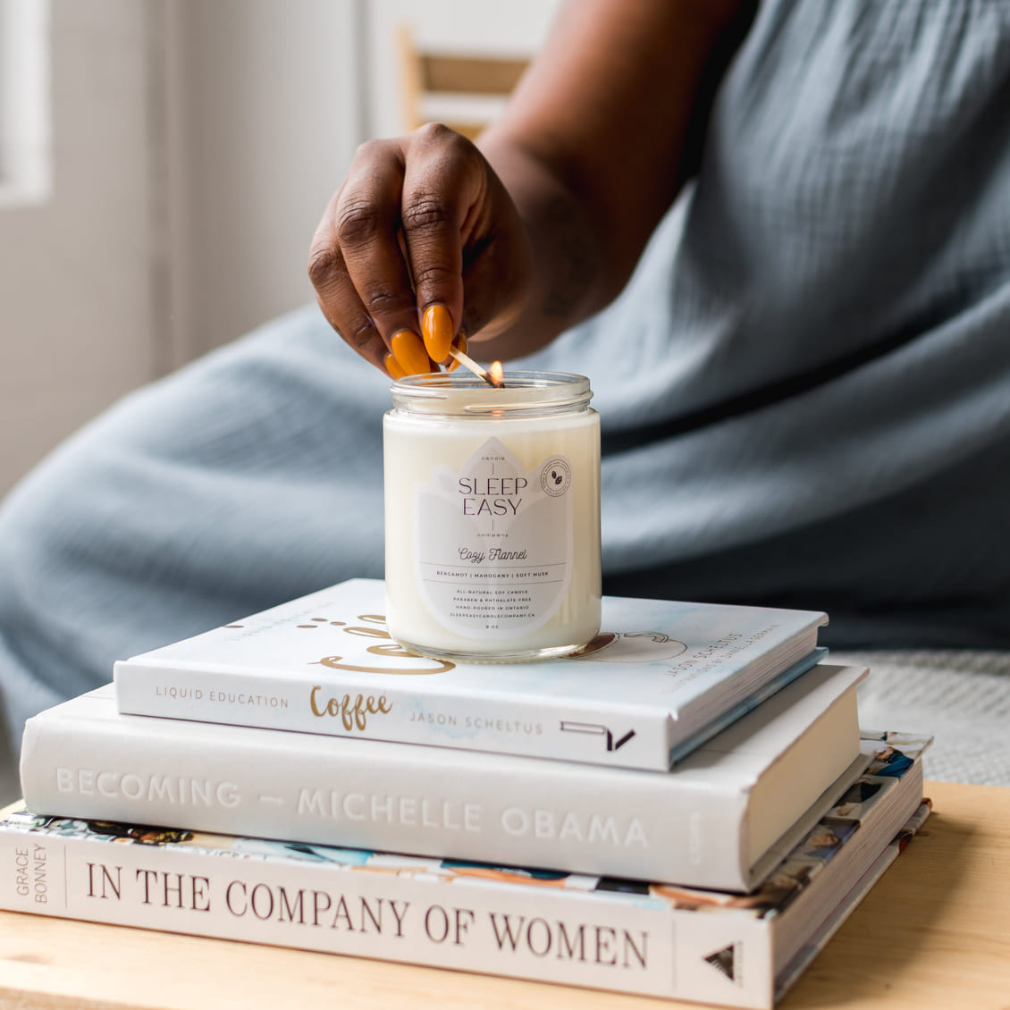 White candle with light blue label is on top of a stack of books. A woman is lighting the candle.  