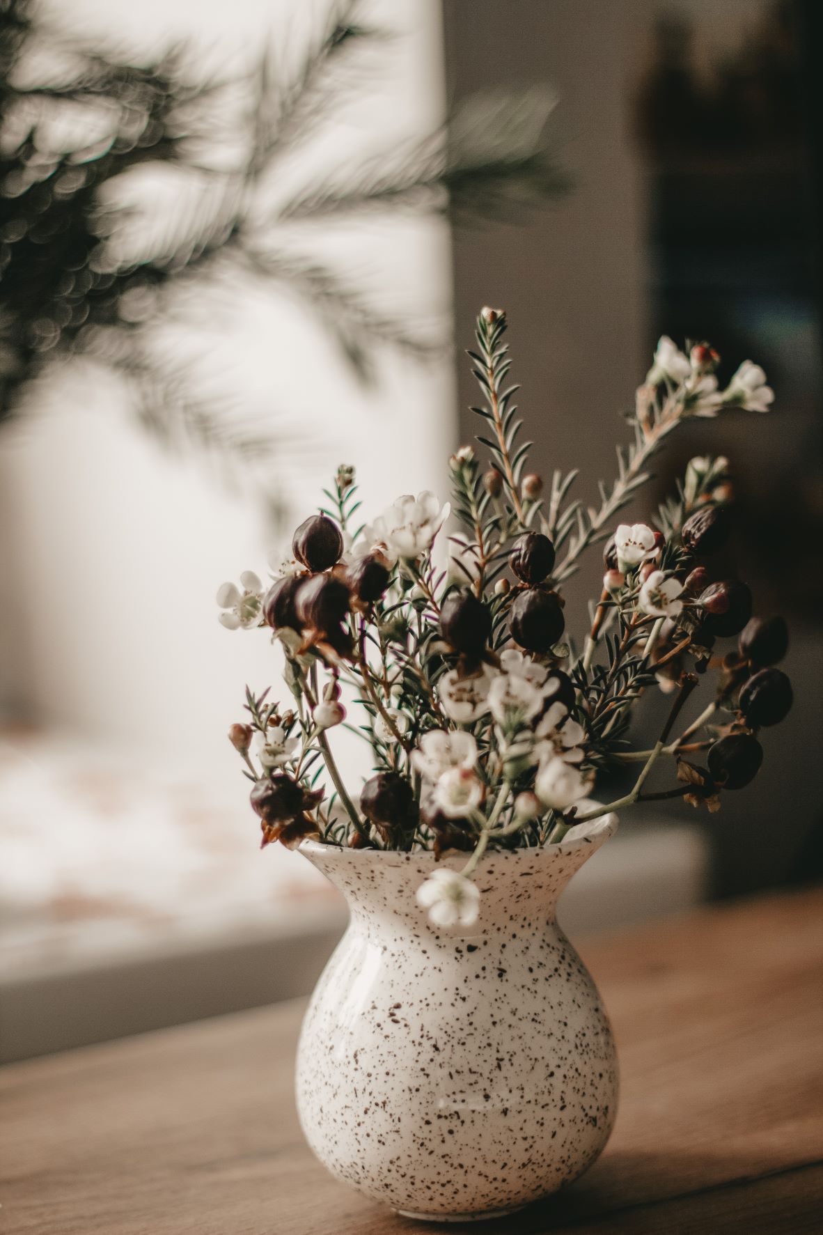 Woody herbs and red berries are displayed in a cream coloured vase on top of a wood table.