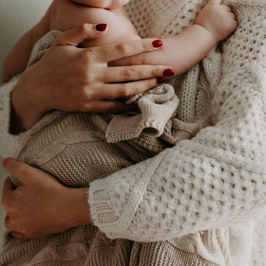 A mother is holding her new born baby in her arms. She is dressed in a warm knitted sweater.